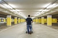 Lonely man walks with a shopping cart in an empty underground parking with yellow concrete columns and neon on the ceiling