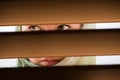 Lonely muslim young woman in a headscarf with frightened eyes looks through the wooden blinds. Concept.