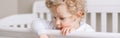 Lonely pensive baby boy toddler standing in crib at home. Sad unhappy thinking baby boy with curly blond hair looking down. Royalty Free Stock Photo