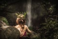 Lonely pensive child girl in isolation in wild forest looking at waterfall with plant wreath on her head as exploring wild nature