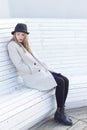 Lonely sad beautiful girl in a black coat and hat, sitting on a white bench cold winter sunny day