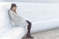 Lonely sad beautiful girl in a black coat and hat, sitting on a white bench cold winter sunny day