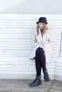 Lonely sad beautiful girl in a black coat and hat, sitting on a white bench cold winter sunny day