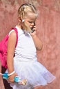 A lonely sad little girl with a backpack and toy in her hand near an old wall in the street was lost and calling on mobile phone Royalty Free Stock Photo