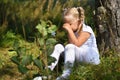 Lonely sad little girl in a white dress and a flower in her hand was lost in the woods, sitting near a tree and crying during day Royalty Free Stock Photo