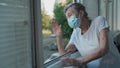 Lonely senior woman in mask stands by window and waves her hand to family who came visit her at hospital. 90 year old Royalty Free Stock Photo