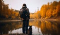Lonely traveler with a pet. Man with backpack stands near the lake and grey cat sit beside him. Yellow autumn trees at backdrop.