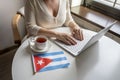 Lonely woman freelancer with flag of Cuba enjoying having breakfast with cup of coffee working on laptop sitting near window