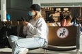 Lonely woman in medical mask and mobile phone in hands sits on chairs with `keep distance` signs in airport waiting room.