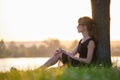 Lonely woman sitting alone on green grass lawn leaning to tree trunk on lake shore on warm evening. Solitude and Royalty Free Stock Photo