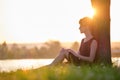 Lonely woman sitting alone on green grass lawn leaning to tree trunk on lake shore on warm evening. Solitude and Royalty Free Stock Photo