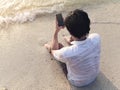 Lonely young Asian man with mobile smart phone sitting on sand of tropical beach.