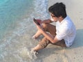 Lonely young Asian man with mobile smart phone sitting on sand of tropical beach. Royalty Free Stock Photo