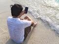 Lonely young Asian man with mobile smart phone sitting on sand of tropical beach. Summer vacations concept. Royalty Free Stock Photo