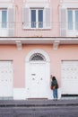 Lonely young female standing in front of a pink and white painted building with open windows