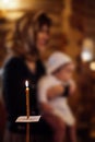Long candle with sheet of paper burning near woman wearing headscarf, holding baby in white clothes in orthodox church.