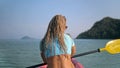 Long curly haired blonde woman in swimsuit rows plastic kayak along azure ocean water at exotic resort close backside