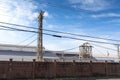 Long dark wall separating a large industrial facility from the street, heavy electrical power lines before a bright blue sky with