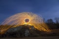 Long exposure shot of man standing on rocky hill spinning steel wool in circle making firework showers of bright yellow glowing sp