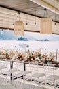 Long festive table with bouquets of flowers stands under a canopy with garlands of light bulbs on the pier