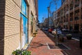 A long gorgeous red brick sidewalk with parked cars along the street and round brown flower pots filled with colorful flowers