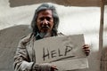 A long-haired Asian man, dirty dressed as a homemaker, holds up a paper placard that reads for help from a passerby because he has