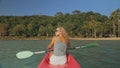 Long haired blonde woman with sunglasses rows bright pink canoe along sea bay water to beach with growing palms. Royalty Free Stock Photo