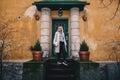 Long haired blonde woman wearing a sand coat with wool sweater, and black jeans, standing on a stairway behind old fashioned house Royalty Free Stock Photo