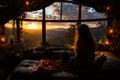 Long-haired female sitting comfortably on a bed near the window. Lady watching the beautiful sunset in the mountains at backdrop