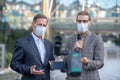 Long-haired male journalist interviewing mature male in front of camera, both wearing masks