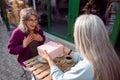 Long haired mature lady presents gift to surprised friend on outdoors cafe terrace