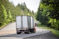 Long haul red big rig semi truck transporting cargo in dry van semi trailer running on the winding highway road with forest on the Royalty Free Stock Photo