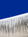 Long icicles hang from the roof of the house against the background of a white brick wall against the blue sky. Royalty Free Stock Photo