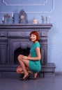 Long legged redhead girl in shiny pantyhose and green minidress posing at fireplace