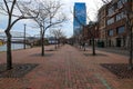 A long red brick sidewalk along the riverfront with rows of bare trees and tall red brick buildings and parked cars