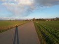 Long shadow photographer on an asphalt road which goes to the village between fields, spring season with cloudy sky