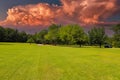 A long smooth winding road surrounded by lush green grass and lush green trees with powerful clouds and red sky Royalty Free Stock Photo