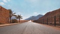 Long straight road with desert hills in distance, brick wall and iron fence on sides. Scenery at the entrance to Wadi Royalty Free Stock Photo