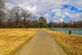 A long winding footpath in the park surrounded by yellow winter grass, bare winter trees, lush green trees, blue sky Royalty Free Stock Photo