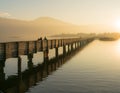 Long wooden boardwalk pier over water in golden evening light with a mountain landscape and people walking silhouette in the backg