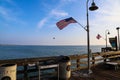A long wooden pier with American flags and curved light posts along the pier with people walking along the pier and blue sky