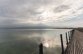 Long wooden boardwalk pier over water with a mountain landscape under an overcast sky