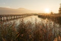 Long wooden boardwalk pier over water in golden evening light with a mountain landscape silhouette in the background and golden ma Royalty Free Stock Photo