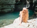 A longhaired girl sits with her head thrown back on the rocky bank of a turquoise river surrounded by canyons