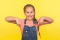 Look, advertise here! Portrait of delighted cute little girl in denim overalls pointing fingers down, showing place below