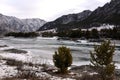 A look at the ice-bound river through low young pines surrounded by high mountains