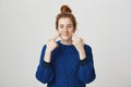 Look at my cute freckles. Indoor shot of calm pretty red-haired female teenager in bun pointing at cheeks with both Royalty Free Stock Photo