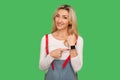 Look at time! Portrait of positive adult woman in stylish overalls pointing at wrist watch and smiling Royalty Free Stock Photo
