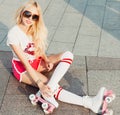 A looker leggy long-haired young blonde woman in a vintage roller skates, sunglasses, T-shirt shorts sitting on road. Summer portr