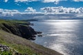 Looking along the rugged cliffs of the Dingle Peninsula, Ireland, bright blue sky with fluffy clouds, sun reflecting off the calm